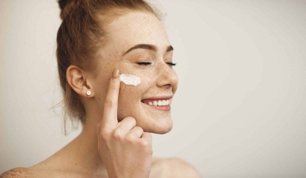 close up of a young female with red hair and freckles applying white cream on her face laughing with closed eyes isolated on white background.